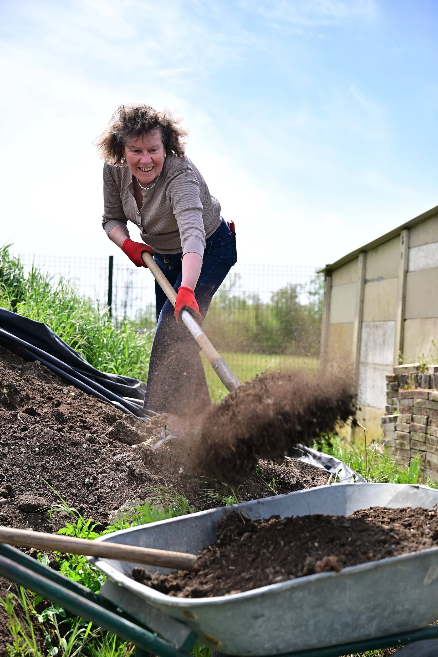 Une femme heureuse en tenue décontractée utilise une pelle pour travailler la terre dans un jardin en plein air. Elle porte des gants rouges et un collier, et se trouve devant un enclos en bois avec un ciel bleu en arrière-plan.