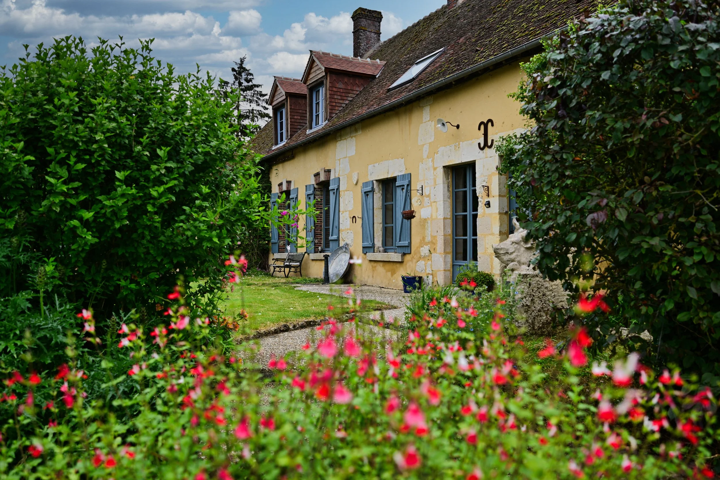 Une maison en pierre avec volets bleus, entourée d'un jardin avec des fleurs roses et des arbustes verts, sous un ciel partiellement nuageux.