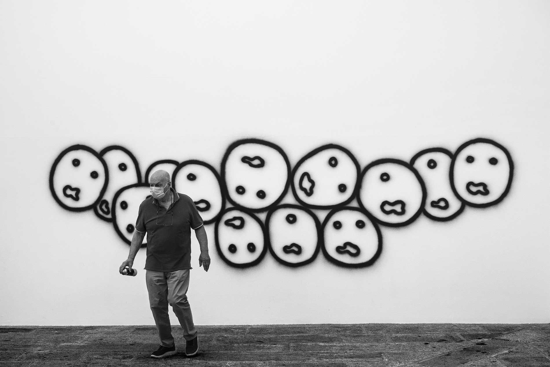 A man wearing a face mask and glasses walking in front of a wall with black outline drawings of various abstract faces.