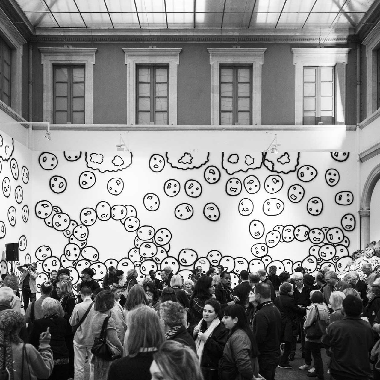 Une foule de personnes dans une galerie d'art avec un mur décoré de dessins de visages au style simple et monochrome.