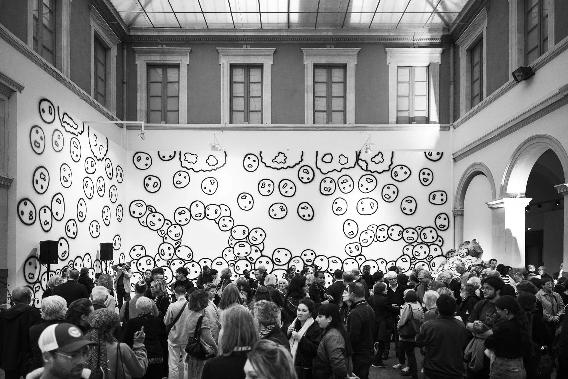 Black and white photo of an art gallery with a large crowd of visitors. The gallery features a wall covered with numerous cartoonish faces and cloud shapes.