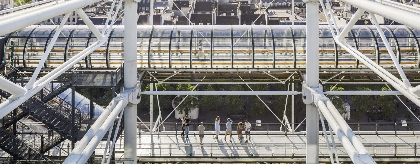Multiple people walking on a glass-covered pedestrian bridge with metal framework.