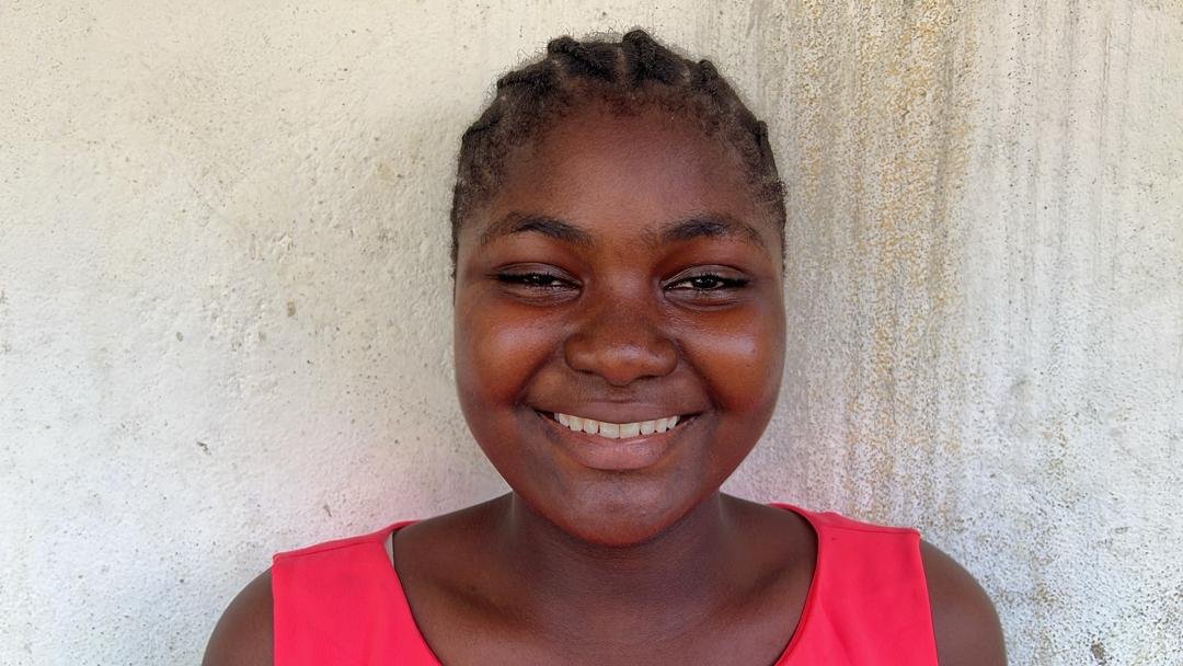 A young woman with short hair and dark skin, smiling at the camera, wearing a pink sleeveless top, standing against a textured light-colored wall.