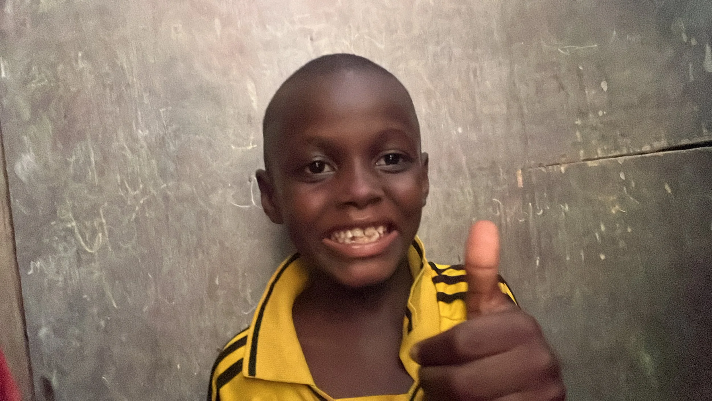 A young boy wearing a yellow and black sports jacket giving a thumbs up and smiling at the camera, standing against a metal wall.