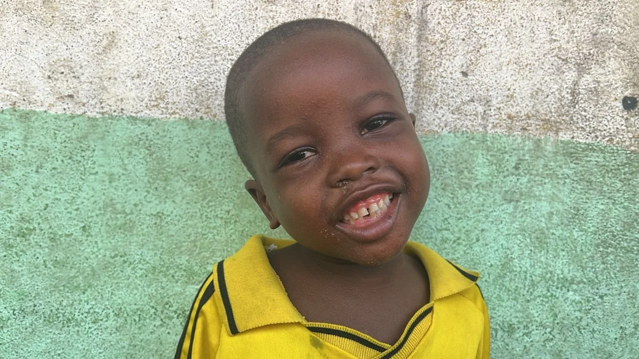 Smiling young boy with short hair, missing front teeth, and dirt around his nose and mouth, wearing a yellow shirt with black stripes, standing against a two-toned wall.