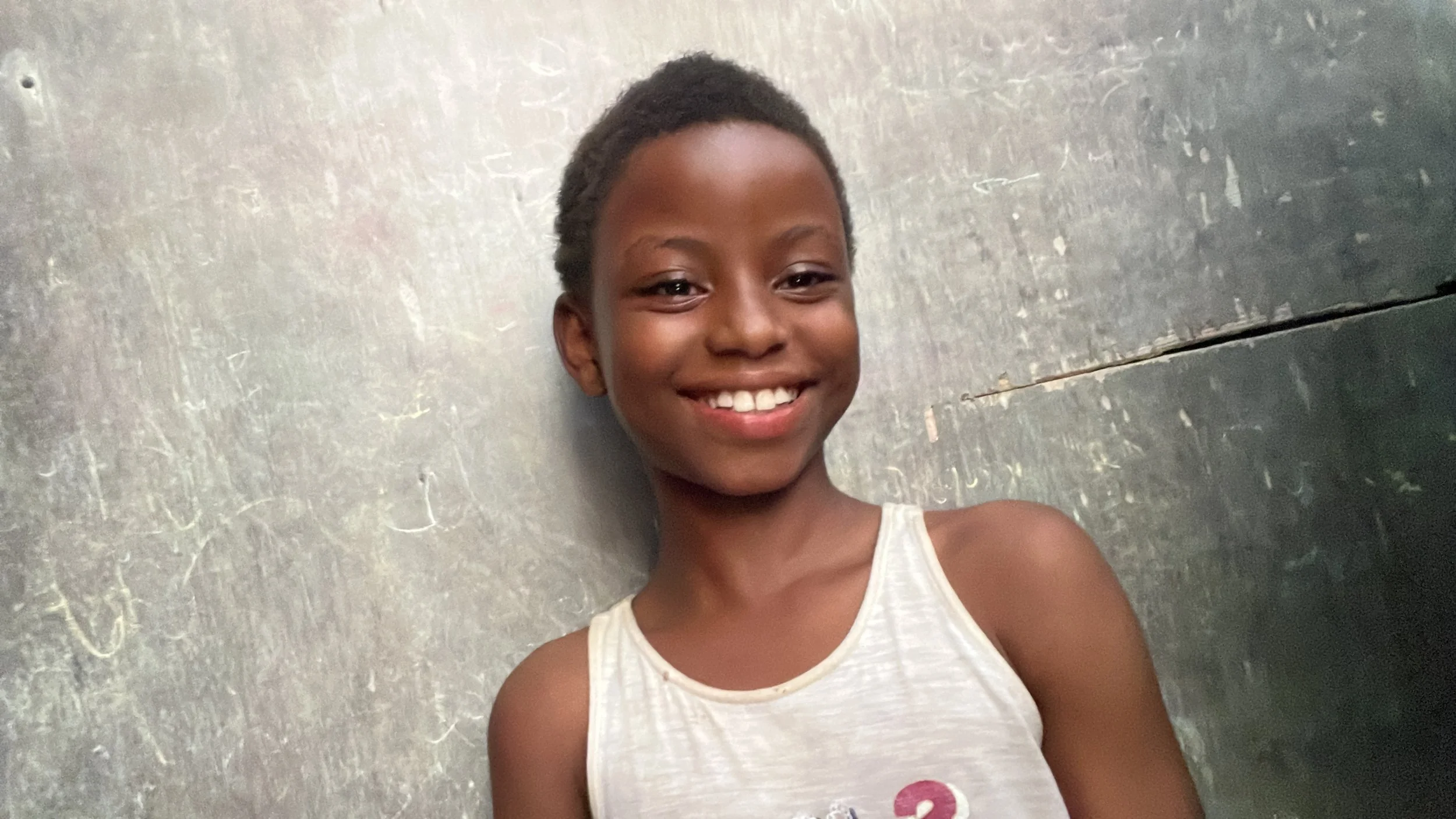 A smiling young girl with short hair and dark skin, wearing a sleeveless white shirt, standing against a textured gray wall.
