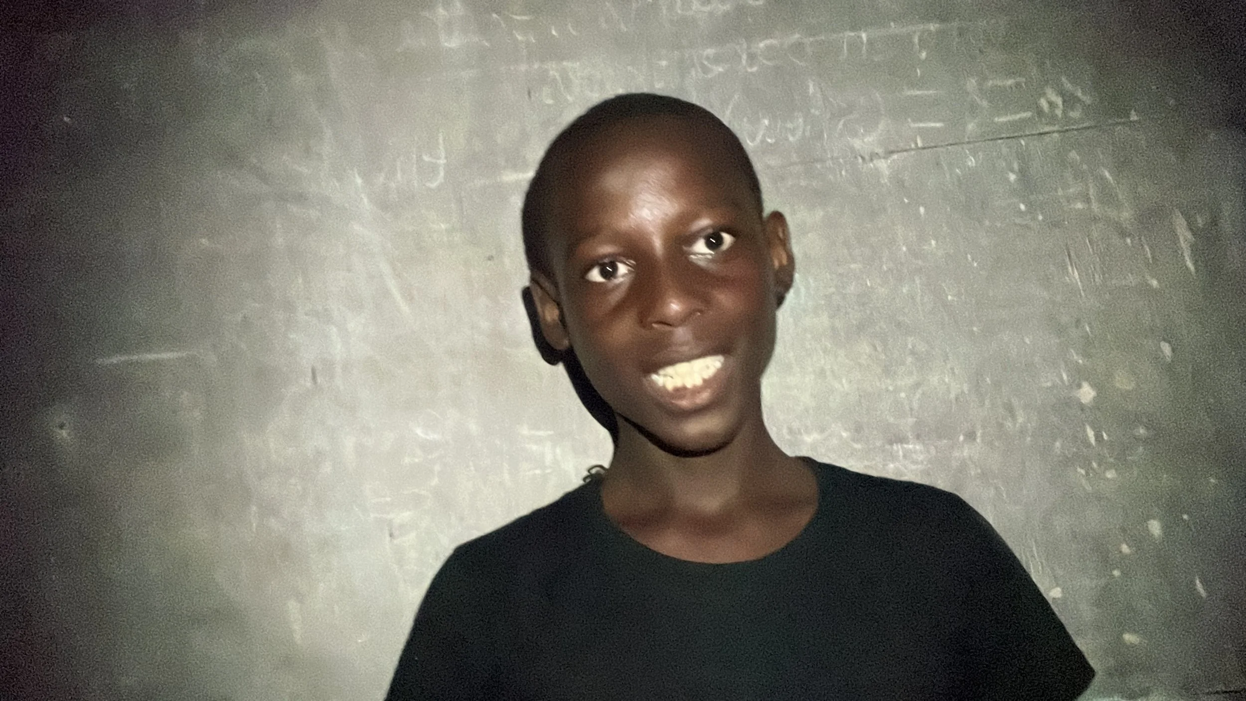 A smiling young boy with dark skin and a shaved head, wearing a black shirt, standing in front of a textured gray background.