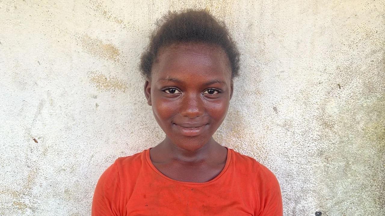 A young girl with dark skin and short, curly hair, smiling and wearing an orange shirt, standing in front of a textured light-colored wall.