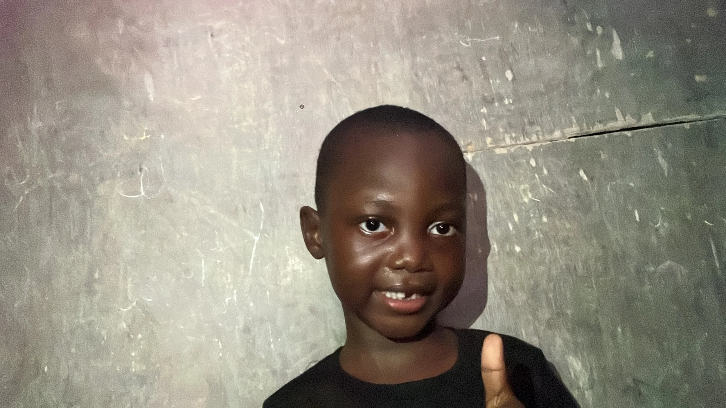 A young boy with dark skin and a short buzz cut hairstyle making a thumbs-up gesture, standing against a metallic wall background.