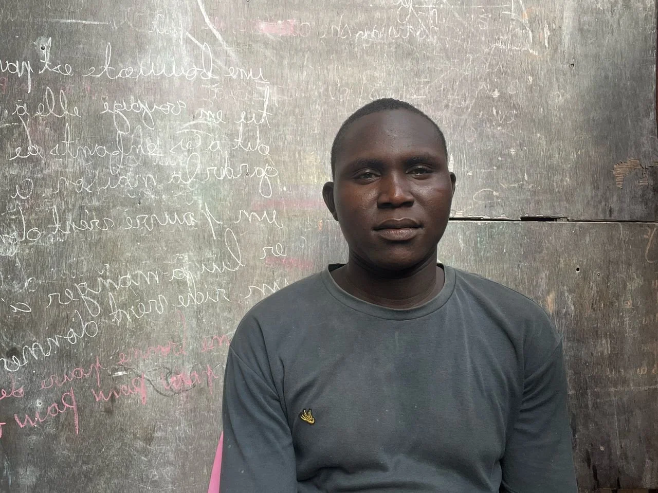 A young man with dark skin and short hair sitting in front of a weathered wooden wall with handwritten text in white and pink chalk.