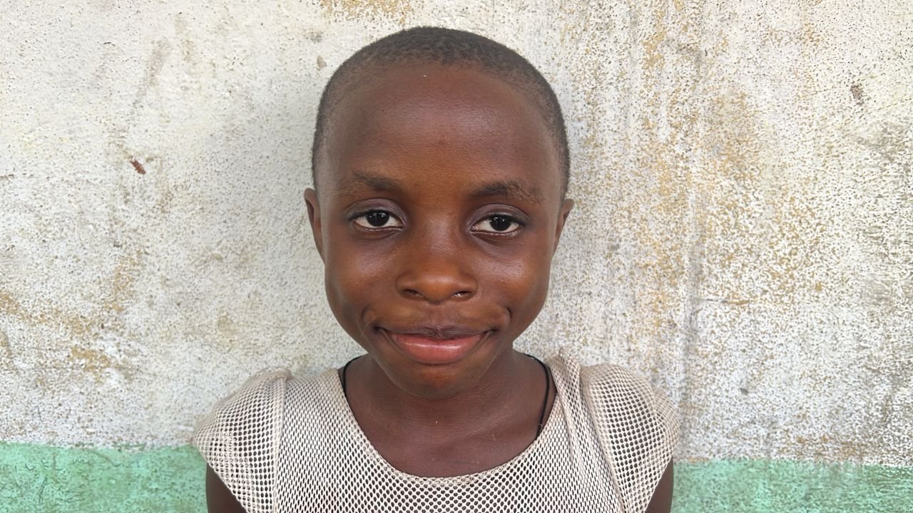A young girl with short hair wearing a mesh top, standing in front of a textured wall with white and green paint.
