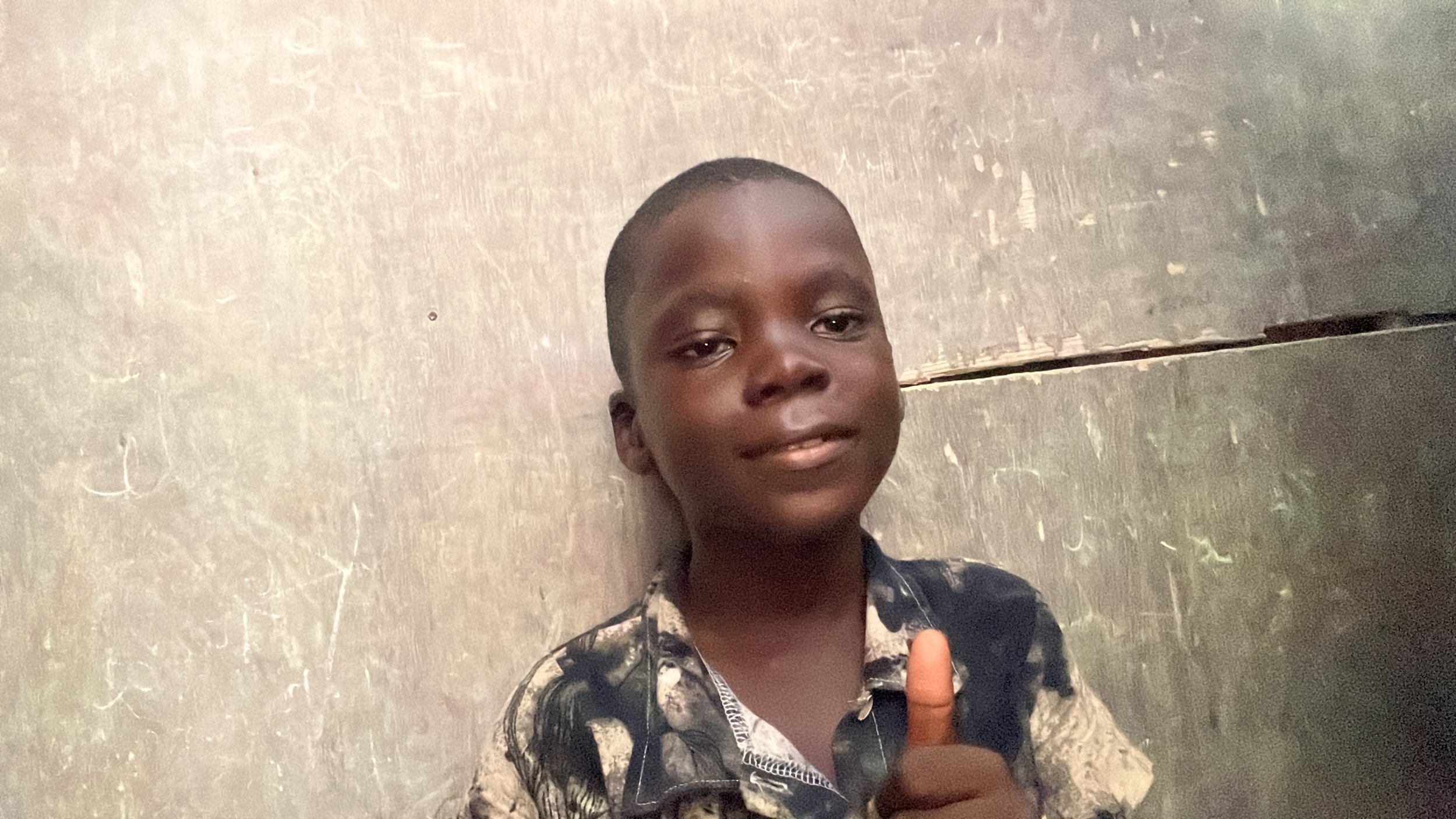 A young boy with short hair giving a thumbs up, wearing a patterned shirt, standing against a plain wall.