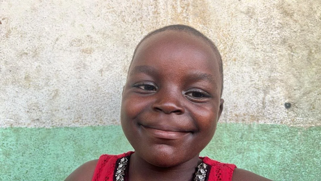 A young boy with a short haircut smiling at the camera, wearing a red sleeveless shirt with embellishments, standing against a textured beige and green wall.