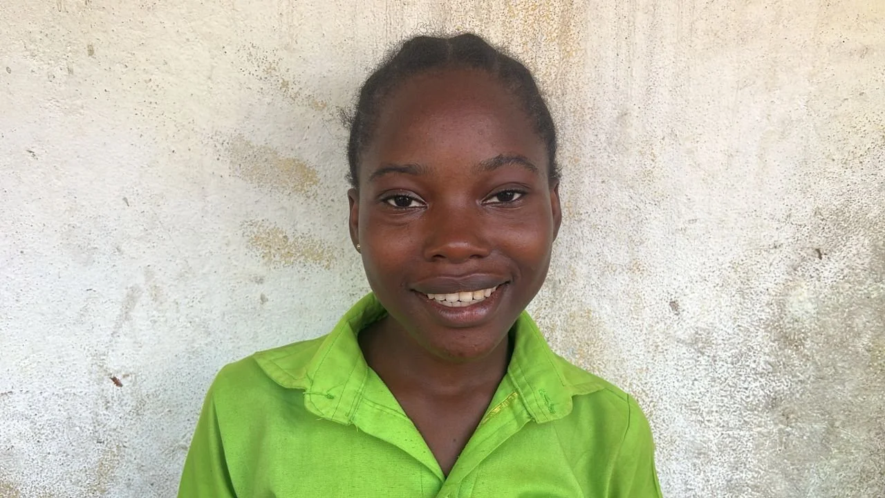A smiling young woman with braided hair wearing a bright green shirt standing against a textured, off-white wall.