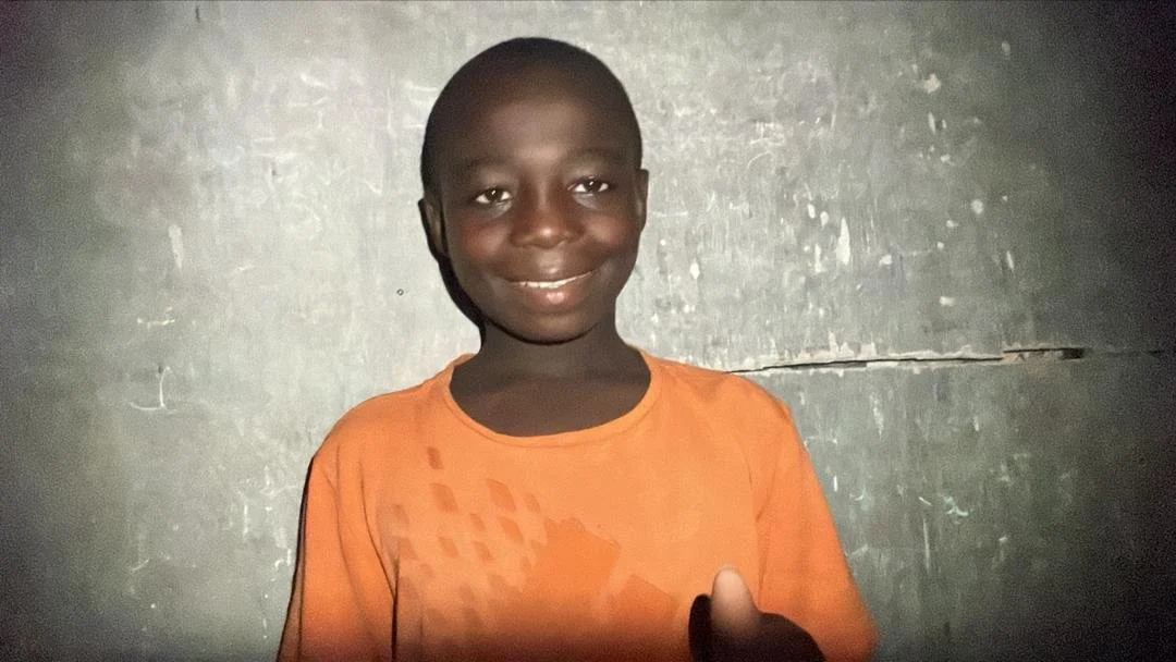 A young boy smiling and standing indoors in front of a plain, worn wall, wearing an orange shirt.