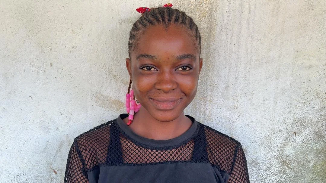 A young girl with braided hair decorated with pink beads and a red bow, wearing a black mesh top over a dark shirt, standing against a light textured wall.