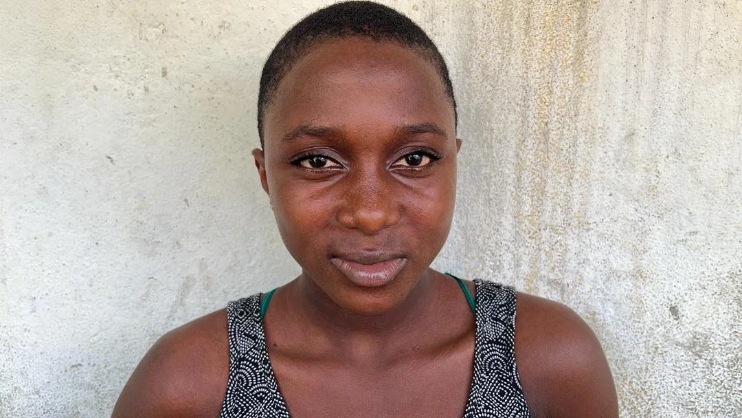 Young woman with short hair wearing a sleeveless top standing against a light-colored textured wall.