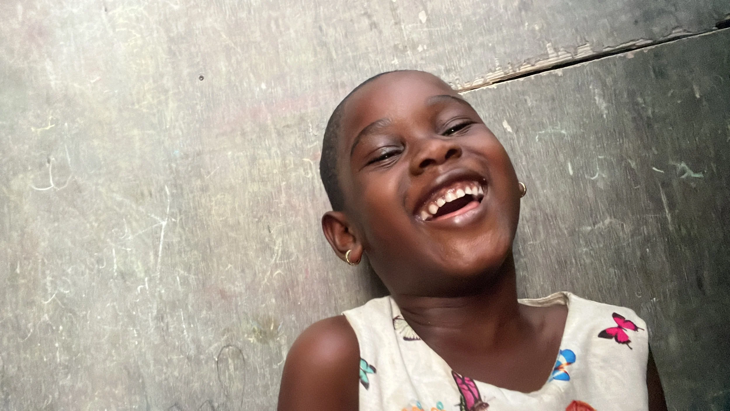 A young girl with short hair and earrings is smiling and laughing, showing her teeth. She is wearing a sleeveless top with butterfly patterns and is standing in front of a weathered, gray wall.
