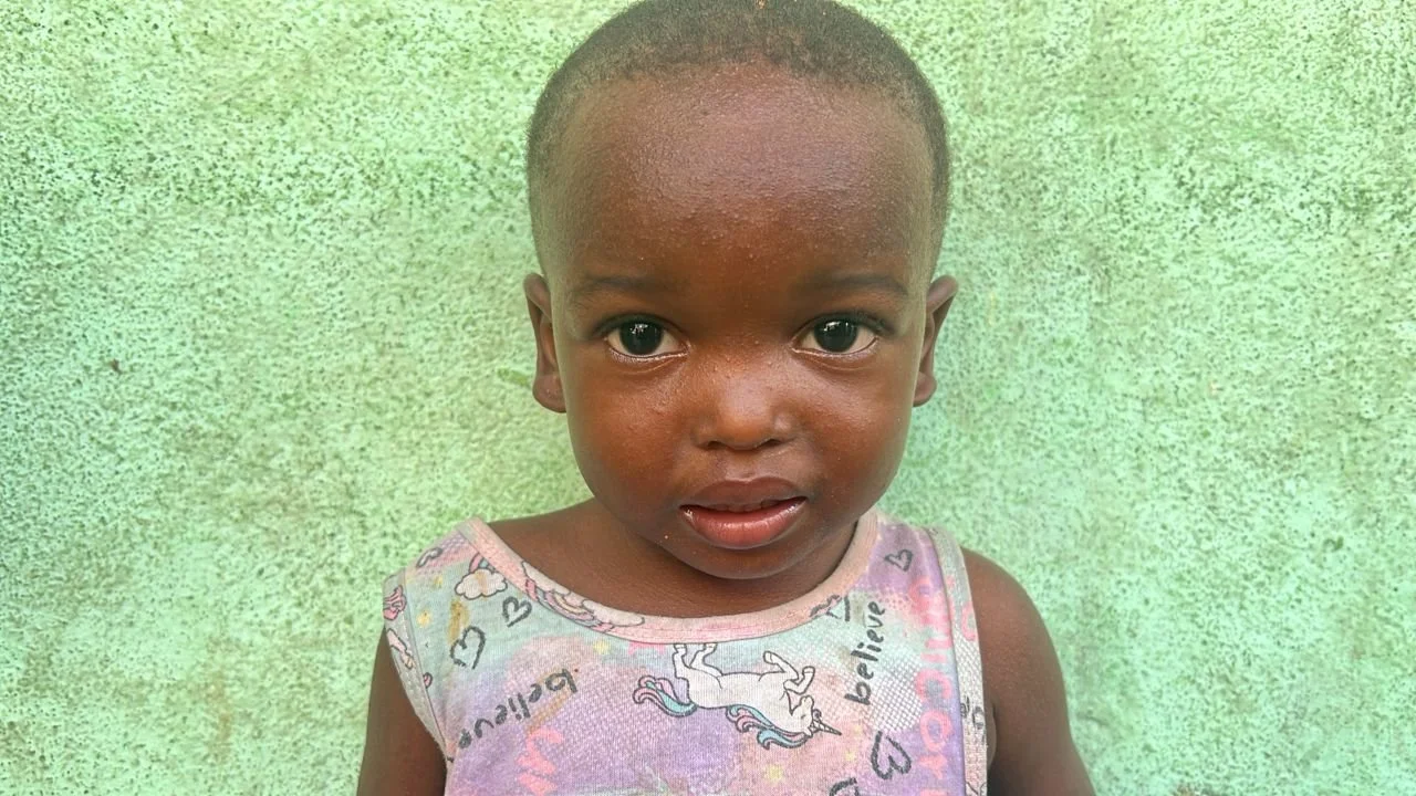 Young child with dark skin and short hair standing against a green wall, wearing a sleeveless shirt with unicorn and heart patterns.