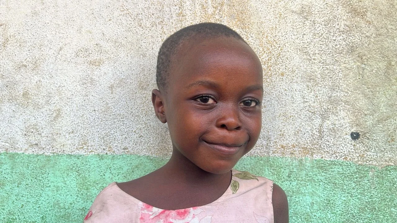 A young girl with a short haircut and a gentle smile, wearing a light-colored shirt with pink and green patterns, stands against a textured wall with a painted green band at the bottom.