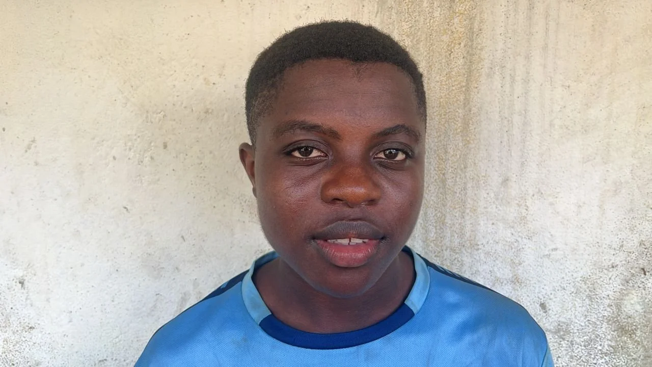 A young person with dark skin and short hair, wearing a blue shirt, standing in front of a textured beige wall.