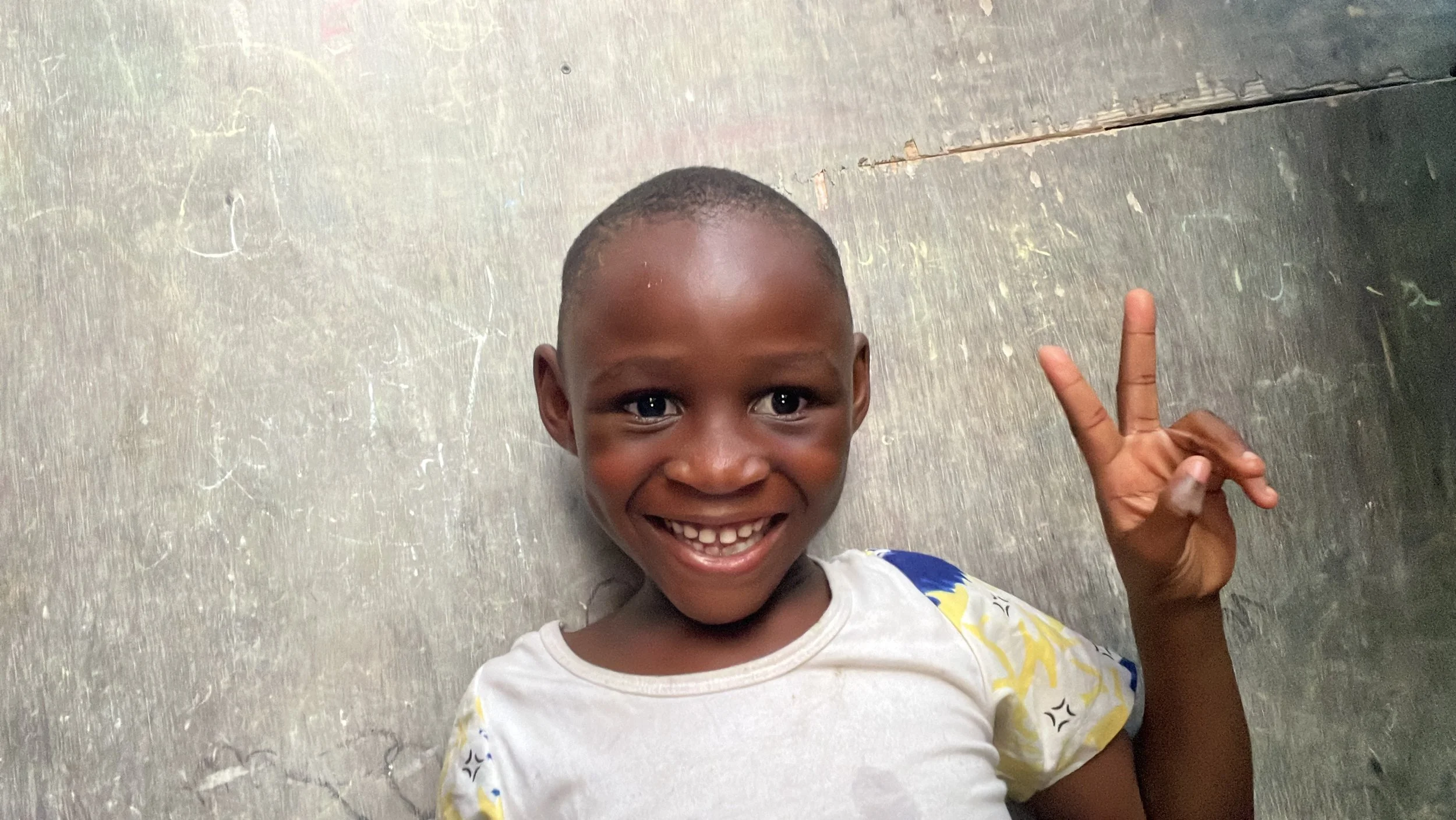 Smiling young African boy lying on the floor, making a peace sign with his right hand, wearing a white shirt with yellow and blue patterns, against a textured gray wall.