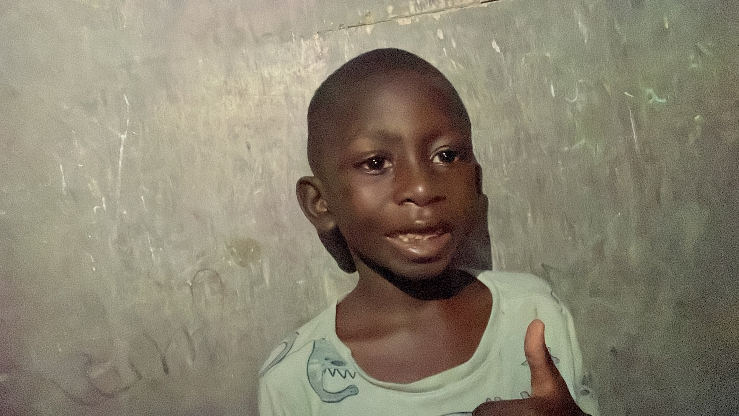 A young boy with dark skin and short hair standing against a textured gray wall, looking to the side with a slight smile and giving a thumbs-up gesture.