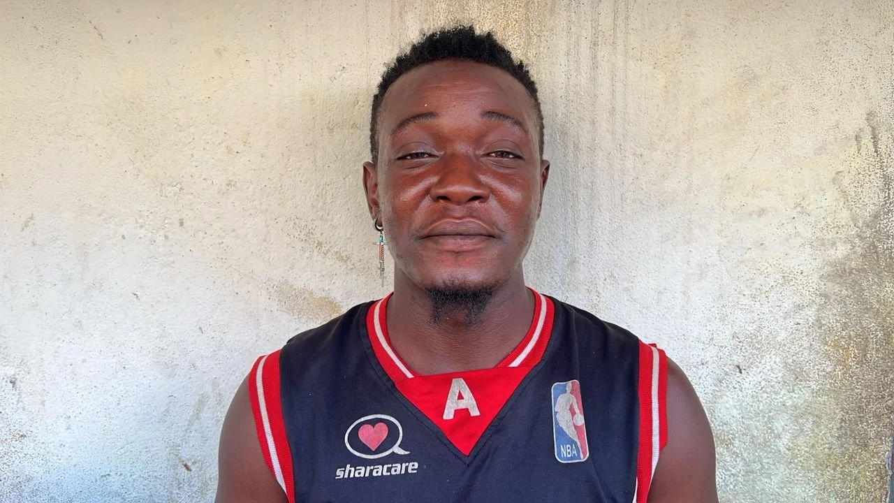 A young man with short curly hair and earrings, wearing a black NBA basketball jersey with red accents, standing in front of a beige textured wall.