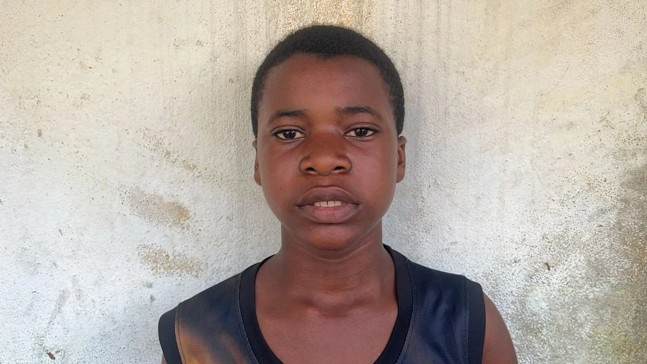 A young boy with short black hair and dark skin, standing in front of a textured off-white wall, wearing a black sleeveless shirt.