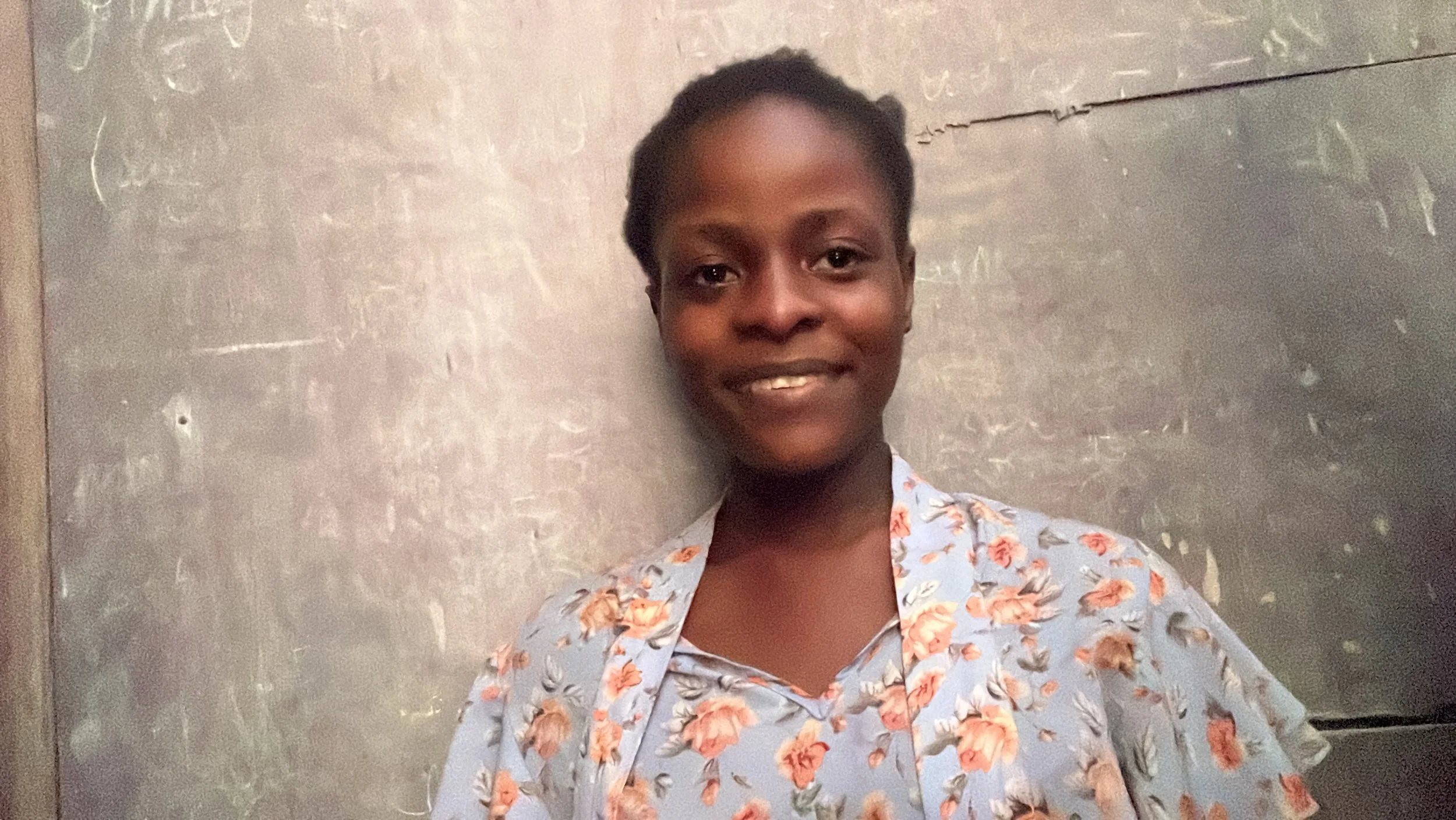 A young woman standing in front of a wall with writing and a crack. She has short hair, dark skin, and is smiling. She is wearing a light blue shirt with a floral pattern.