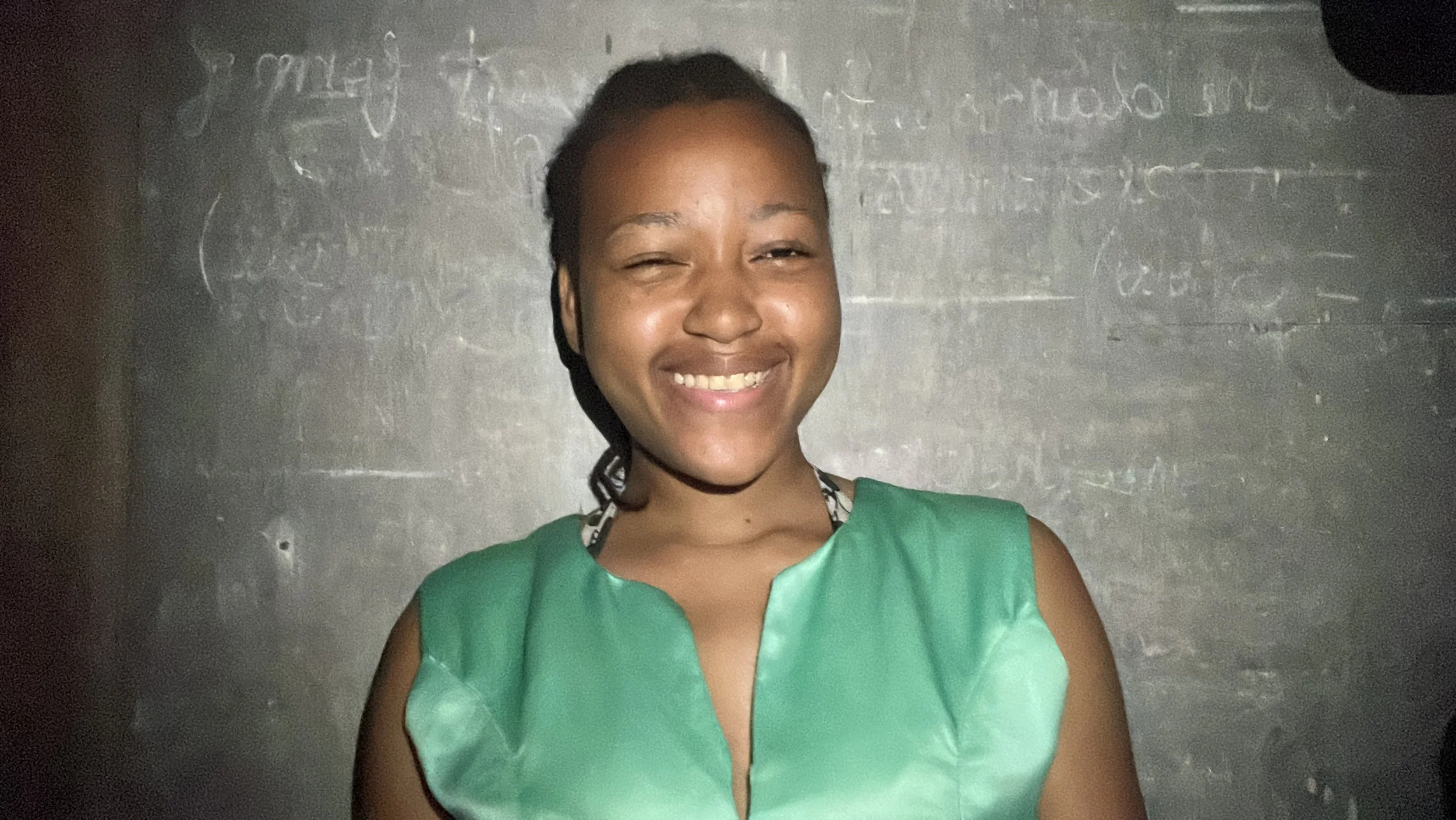 Smiling woman with dark hair tied back, wearing a green top, standing in front of a gray wall with writing.
