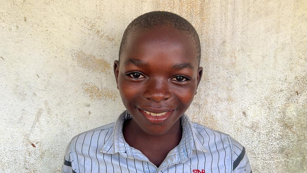 A young boy with a bright smile, wearing a striped shirt, standing against a textured off-white wall.