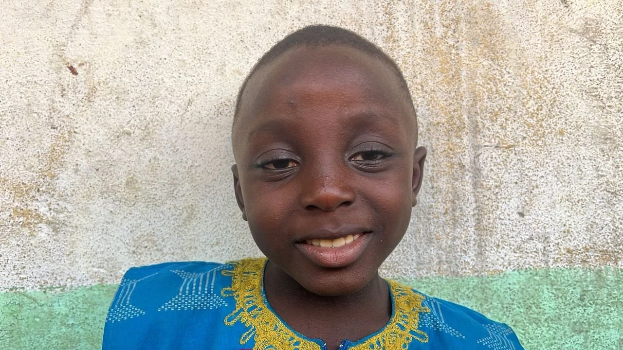 A young boy smiling, wearing a blue shirt with gold embroidery, standing outdoors in front of a textured wall.