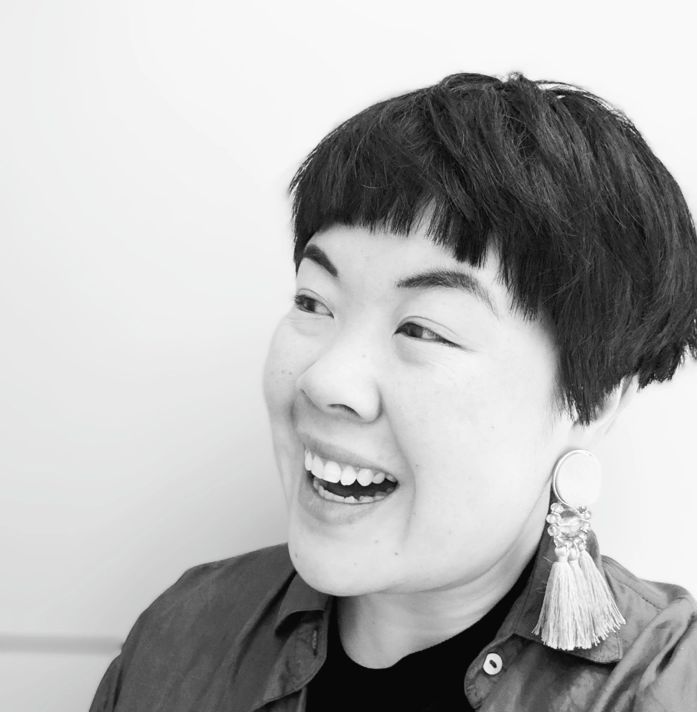 A woman with short, dark hair, smiling and wearing a large tassel earring, in a close-up black and white portrait against a plain background.