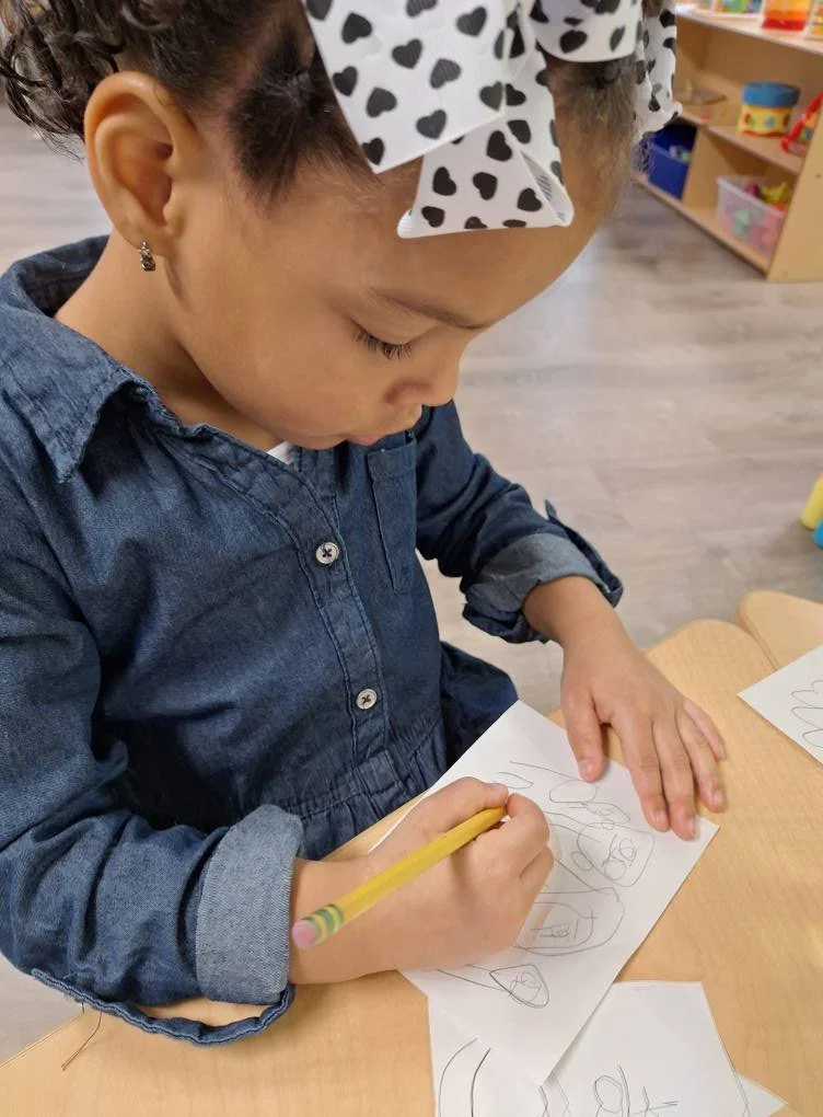 A young girl with dark curly hair tied with a large bow with black hearts, wearing a denim dress, is drawing with a yellow pencil on a piece of paper.