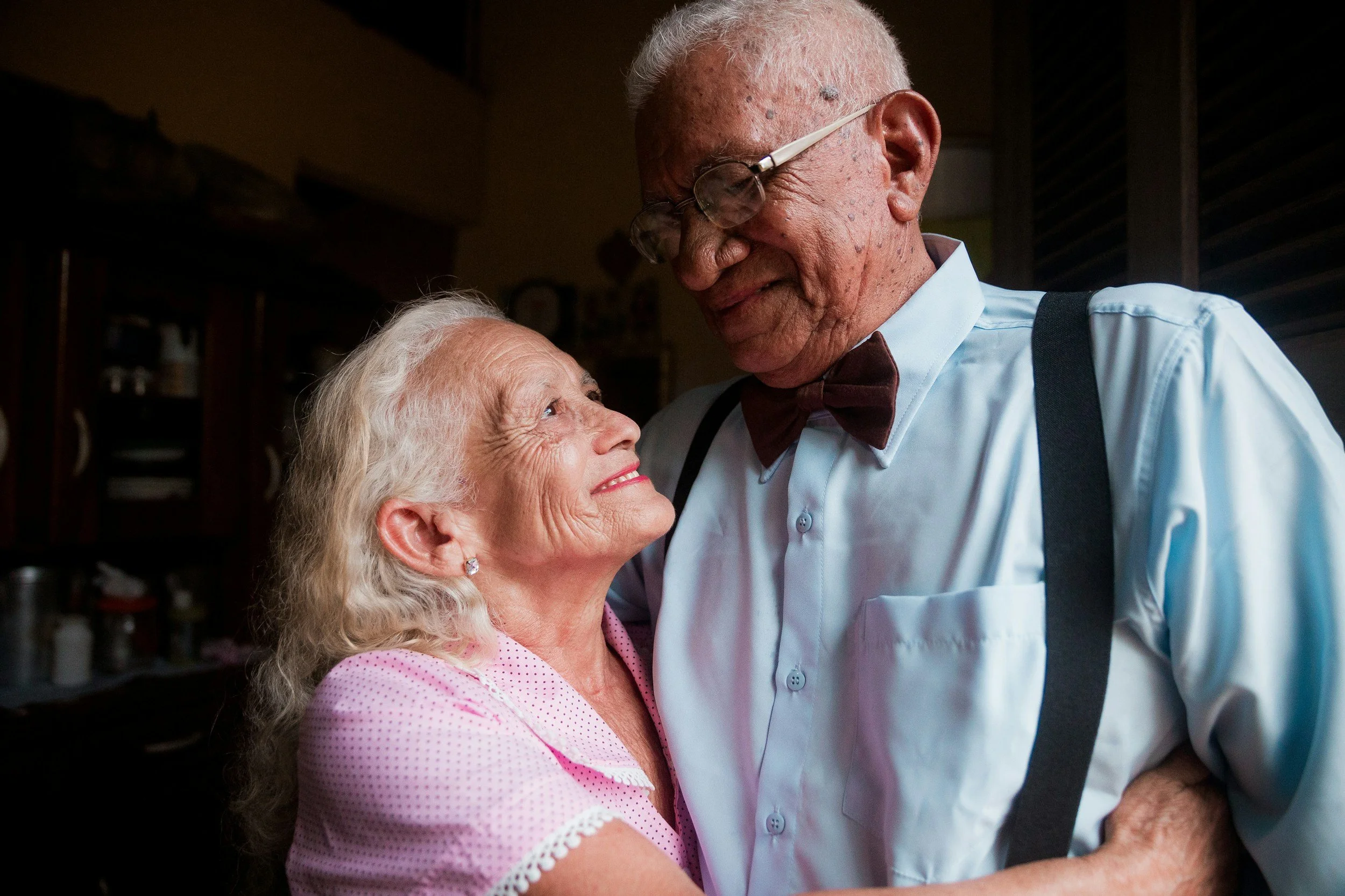 An elderly woman with white hair and a pink dress looking up at an elderly man with white hair, glasses, a white shirt, and a bow tie, who is smiling at her in a warmly lit room.