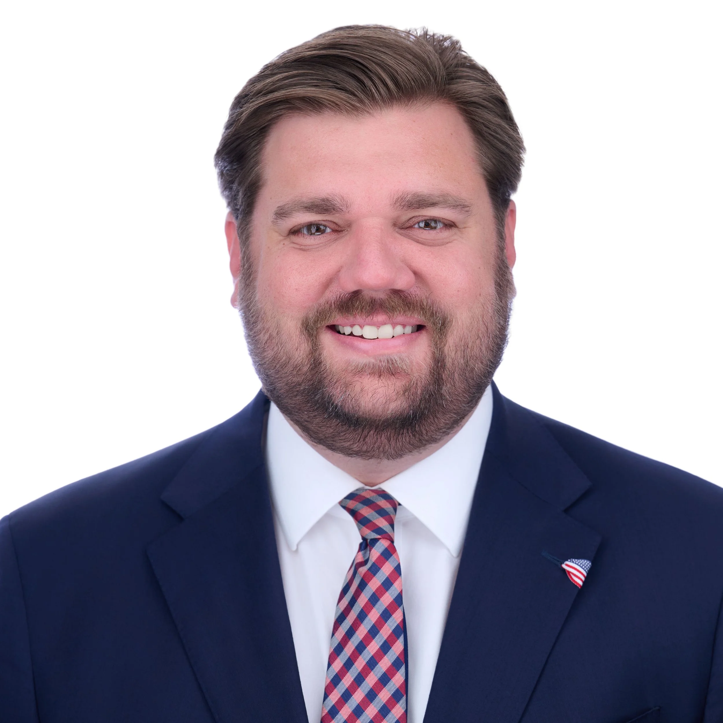Close-up of a smiling man with light brown hair, wearing a dark blue suit, white shirt, and a red, white, and blue plaid tie. He has a small American flag lapel pin on his suit.