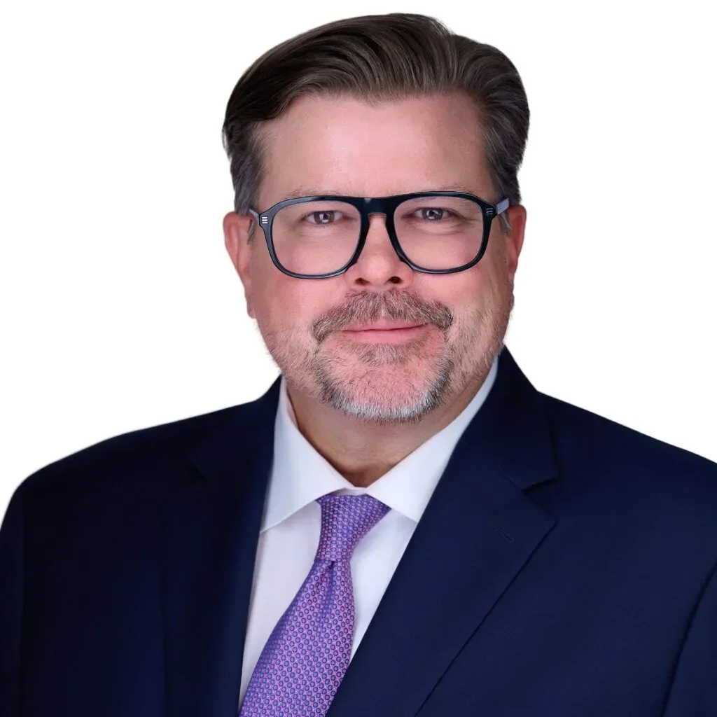 Professional headshot of an older man with glasses, gray hair, and a beard, wearing a dark suit, white shirt, and purple tie, against a white background.