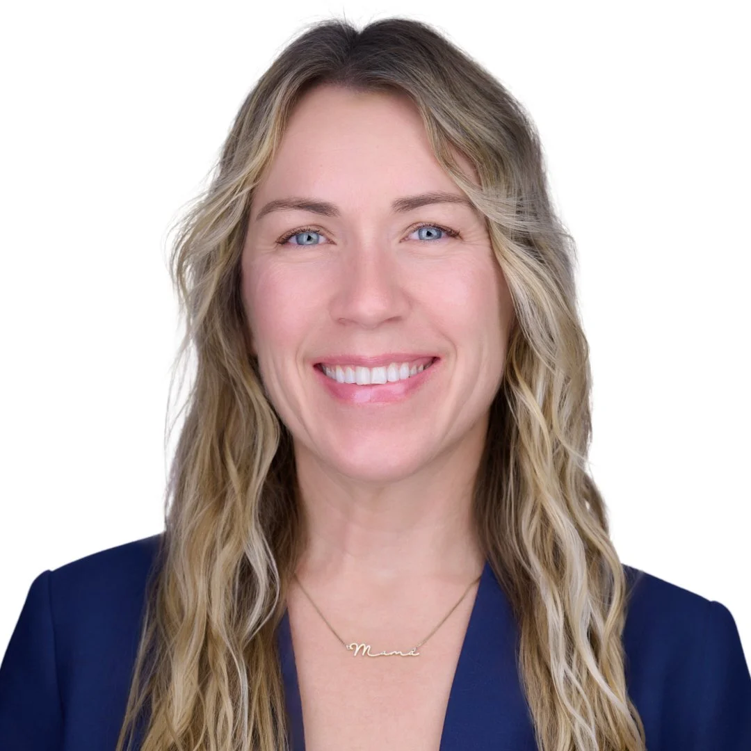 A woman with long, wavy blonde hair, blue eyes, and a friendly smile, wearing a navy blazer and a gold necklace that says 'Mom' in cursive, against a white background.