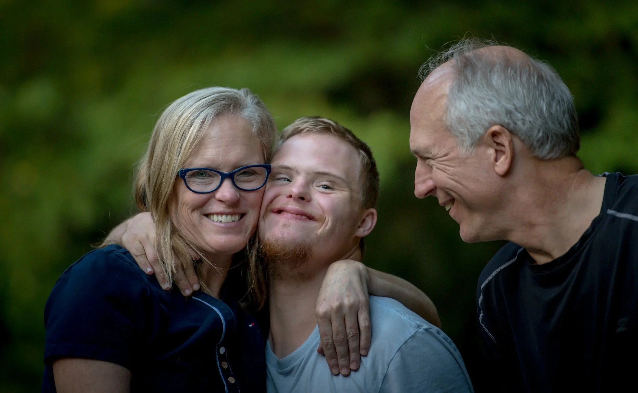 A family of three, a woman with glasses, a young man, and an older man, smiling and hugging each other outdoors with a green background.