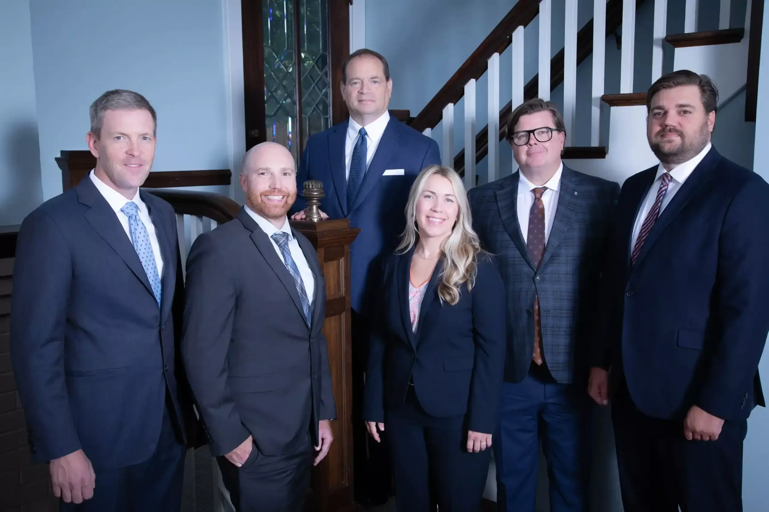 Seven professionally dressed individuals pose together in a formal setting, with a staircase and decorative wall in the background.