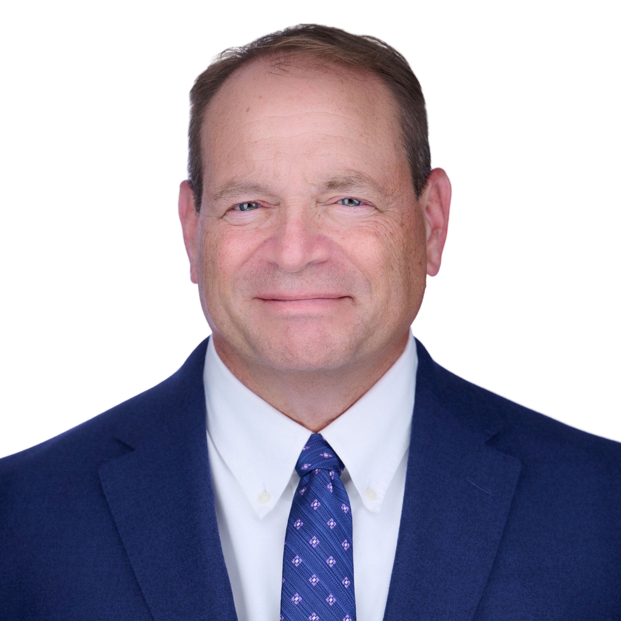 Close-up of a man wearing a dark suit, white shirt, and purple patterned tie, with a friendly expression.