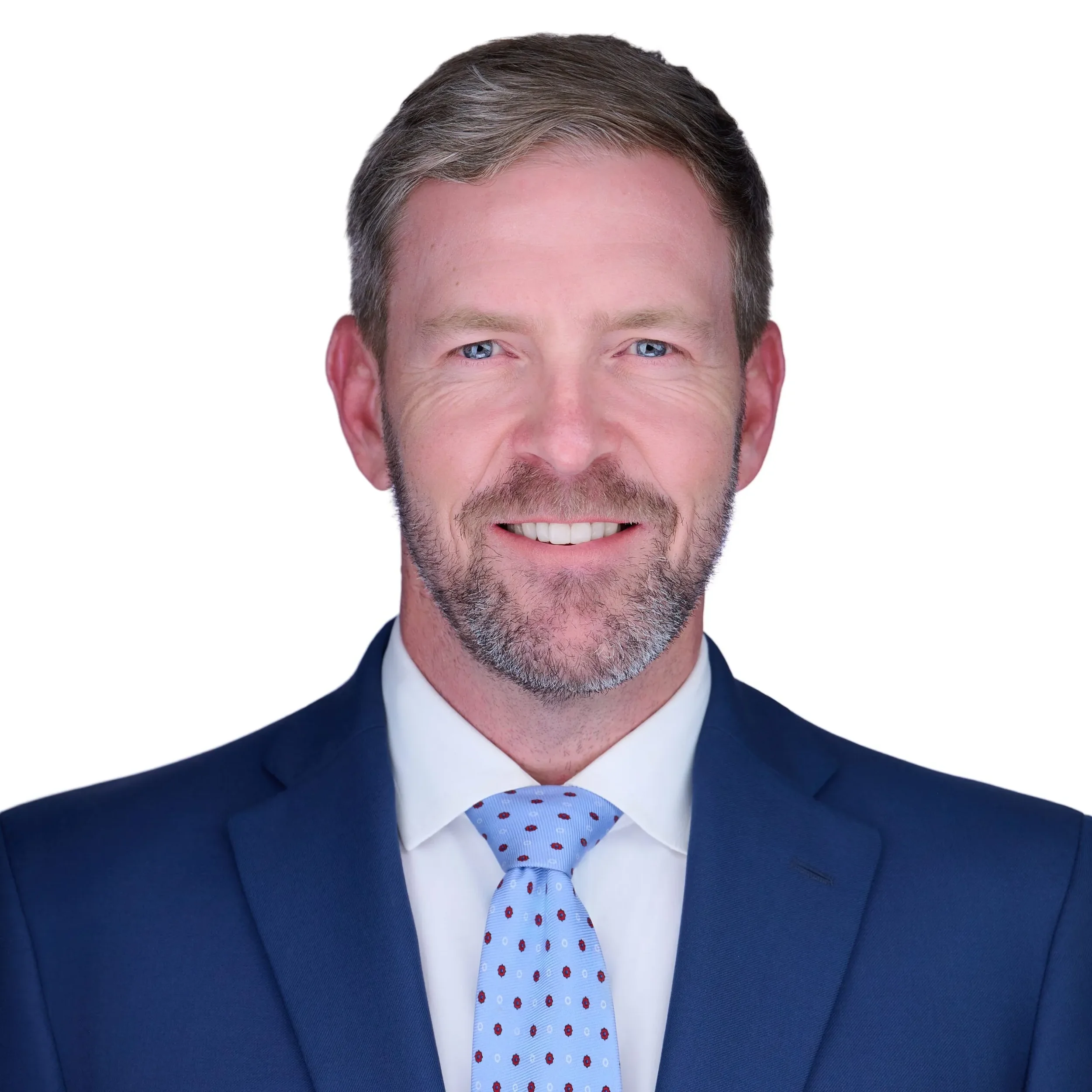 Close-up of a smiling man wearing a blue suit, white shirt, and a light blue tie with red polka dots, with a neatly styled haircut and visible facial hair.