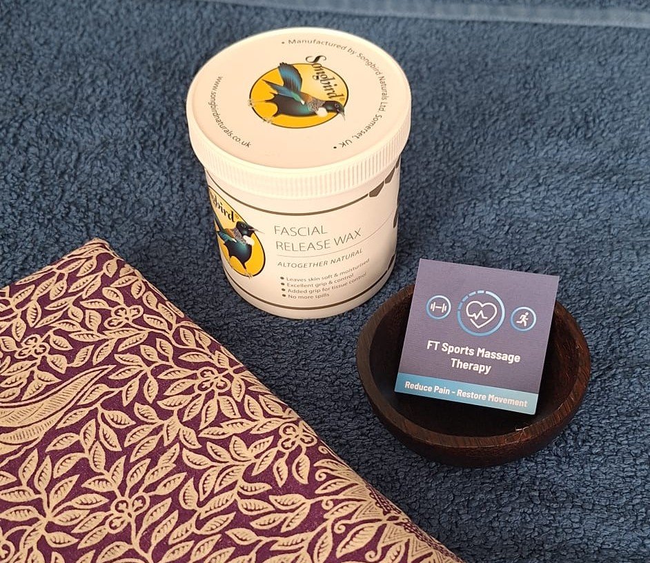 Tub of sports massage wax with bowl with a business card in it on top of a massage bed with blue towels and a purple sarong.