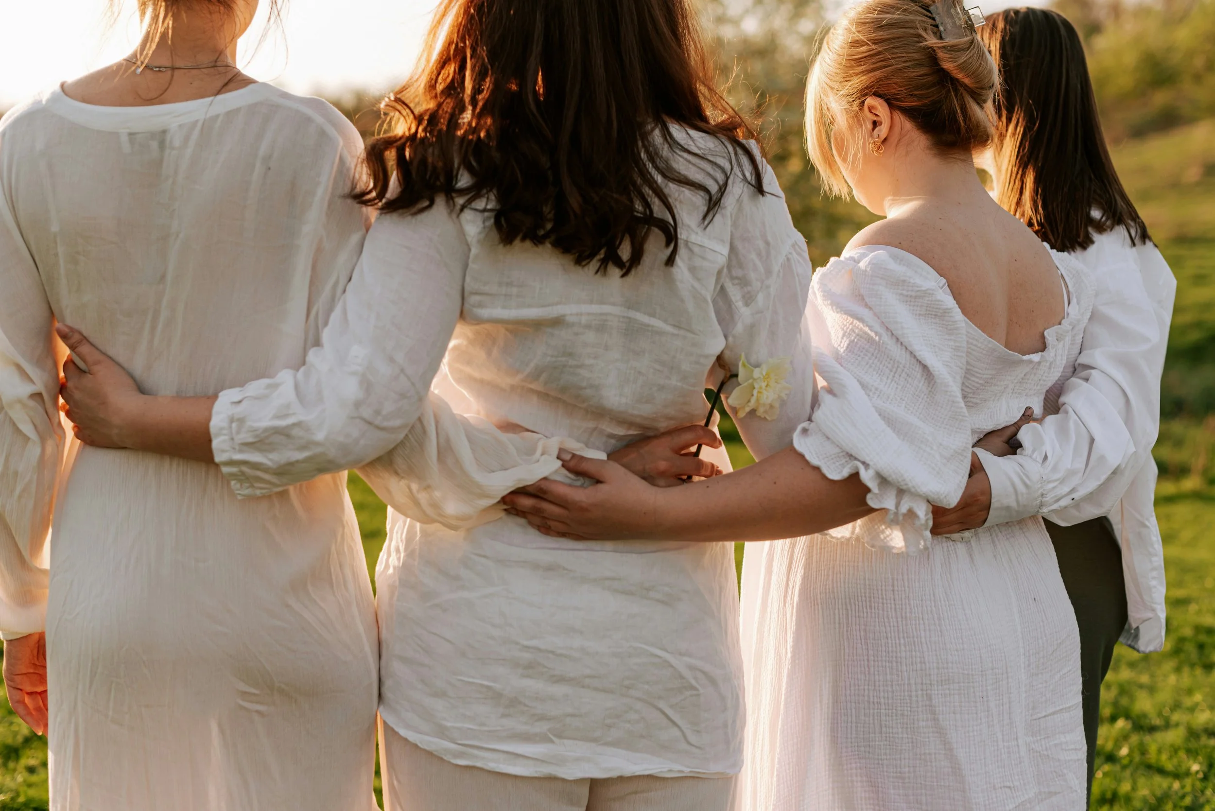 A group of women standing outdoors in white clothing, holding each other and a yellow flower, during a sunset.