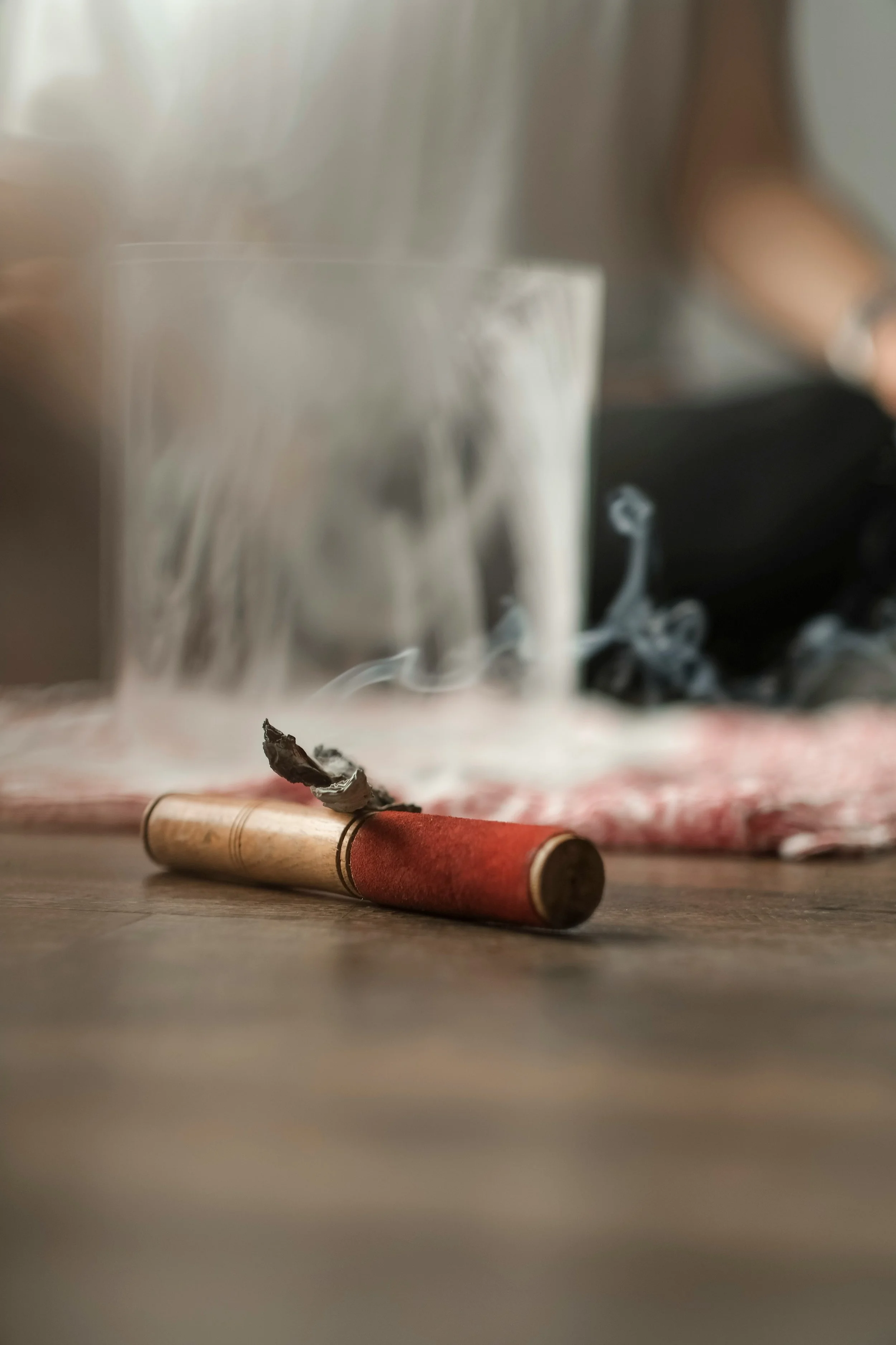 A burnt cigarette lying on the floor with smoke curling from its tip, a glass of milk with milk spilling out, and a blurred background.