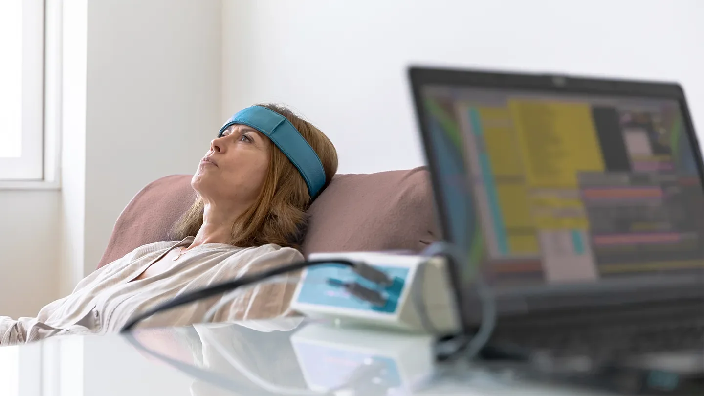 Woman lying on couch with an EEG headband, near a laptop with colorful charts