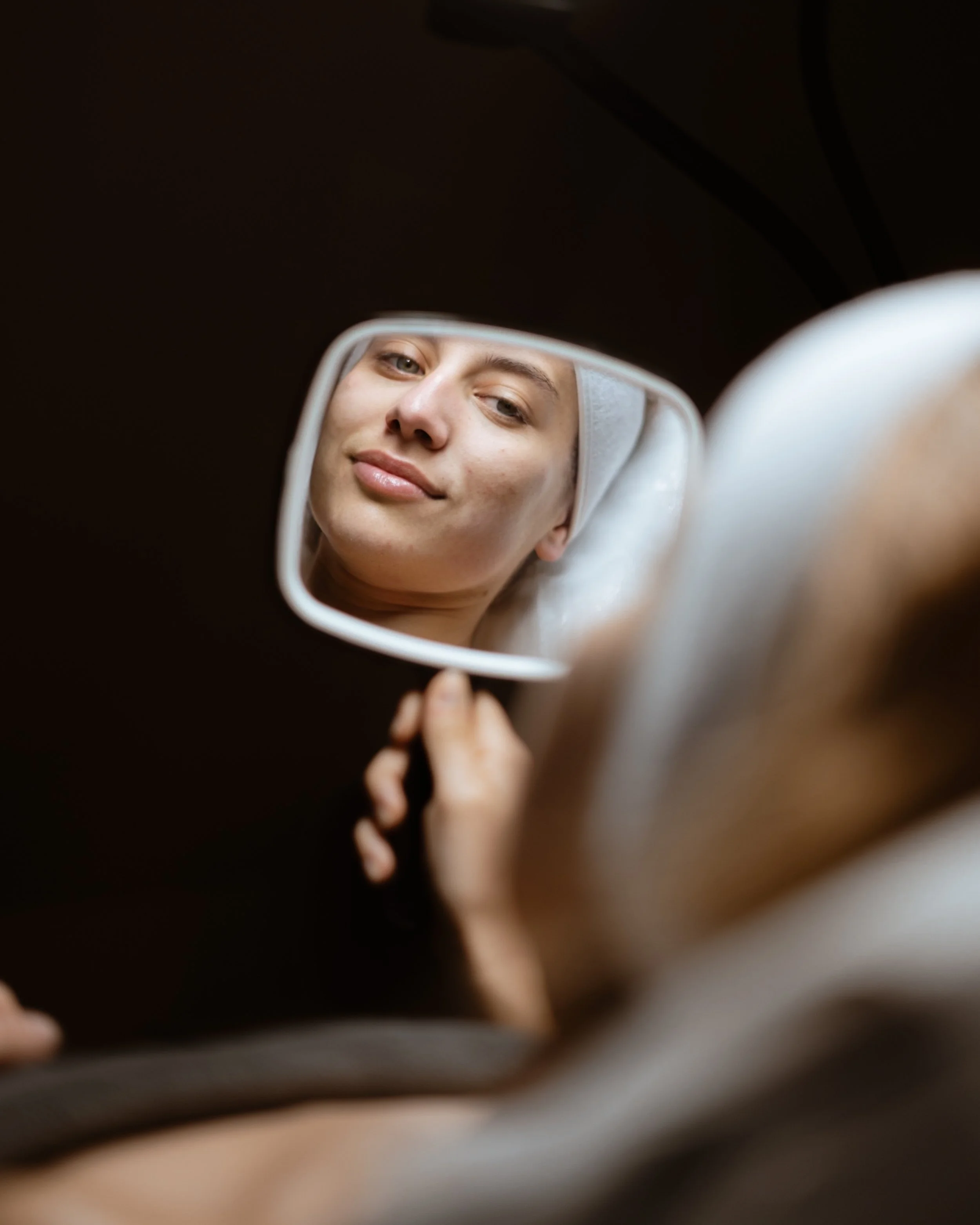 Close-up of a woman lying in a spa, looking into a handheld mirror, with a towel wrapped around her head.