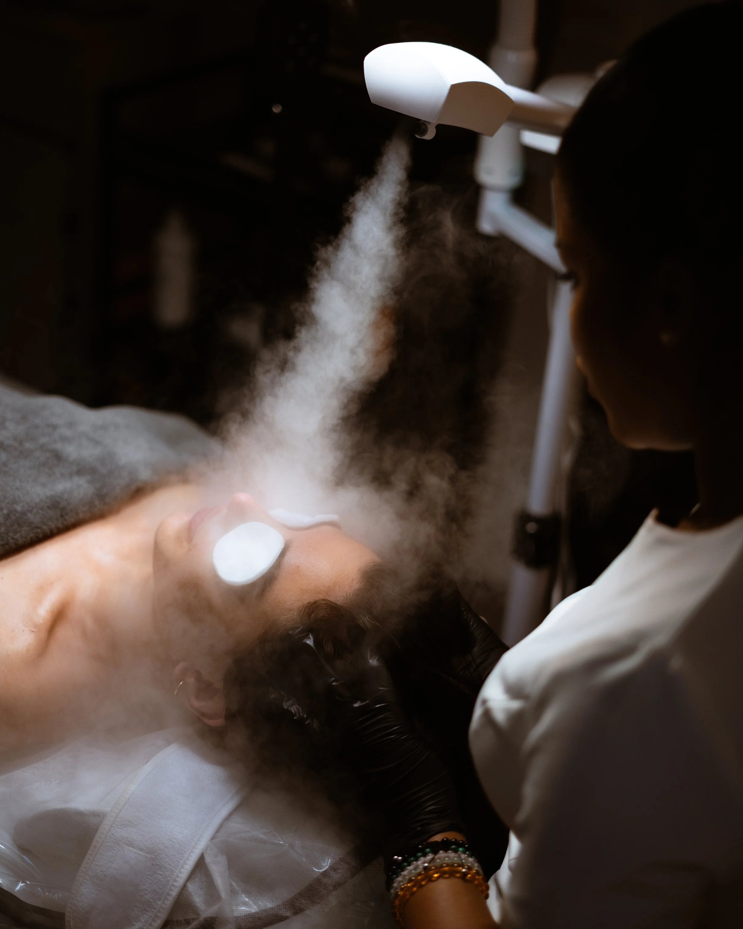 A woman lying on a treatment bed with her eyes closed, receiving a skincare treatment while a technician applies a device emitting steam or mist near her face.