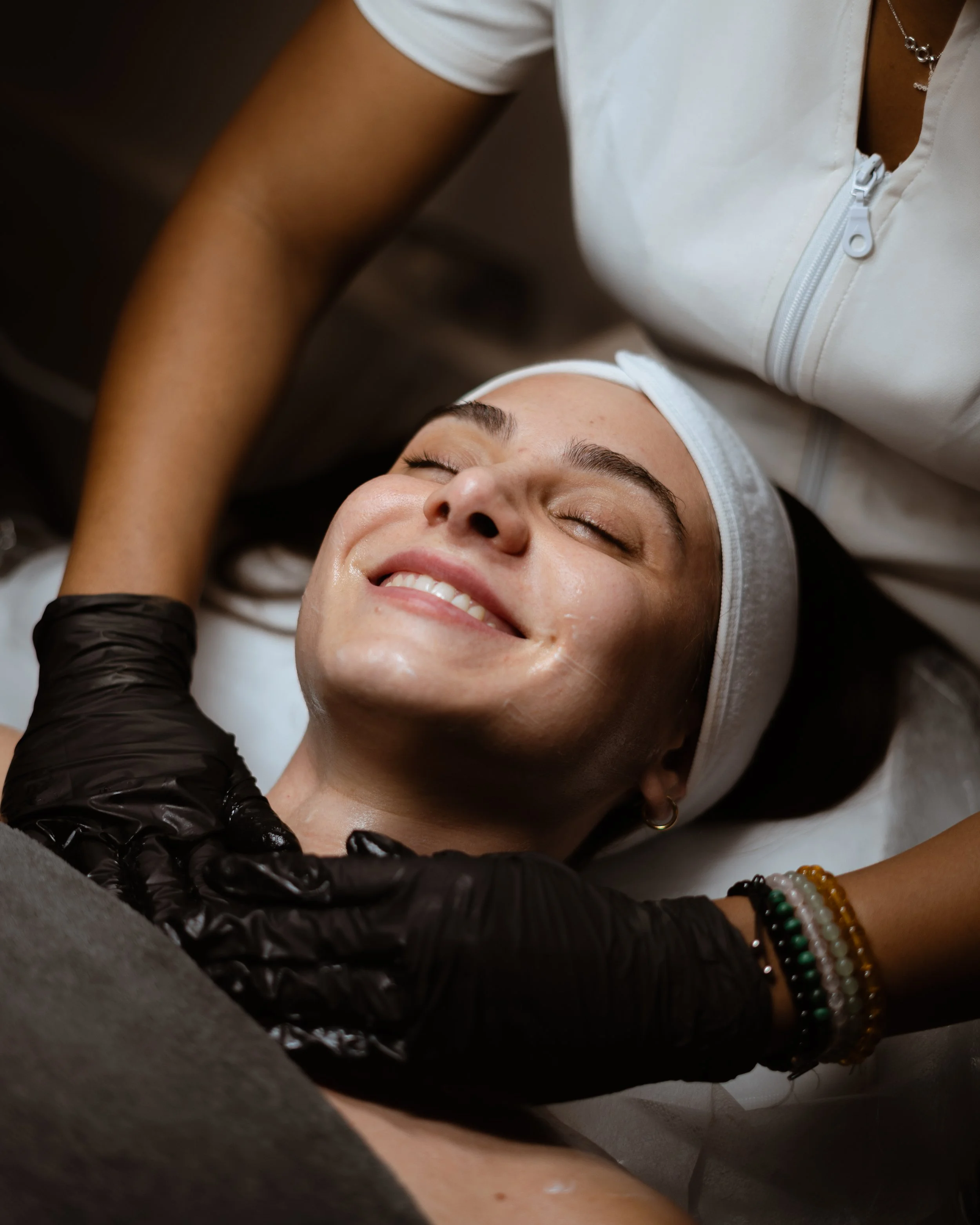 A woman lying on a treatment bed with her eyes closed and smiling, receiving a facial treatment from a professional wearing black gloves.